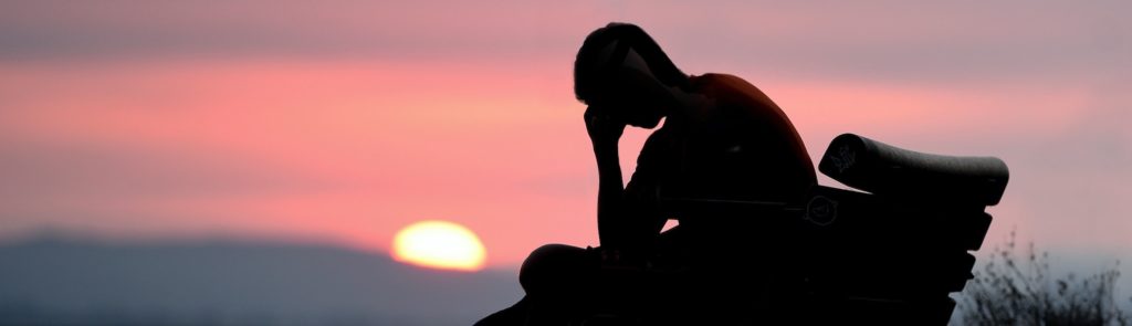 silhouette of a figure seated on a bench alone with his or her head leaning on a hand.